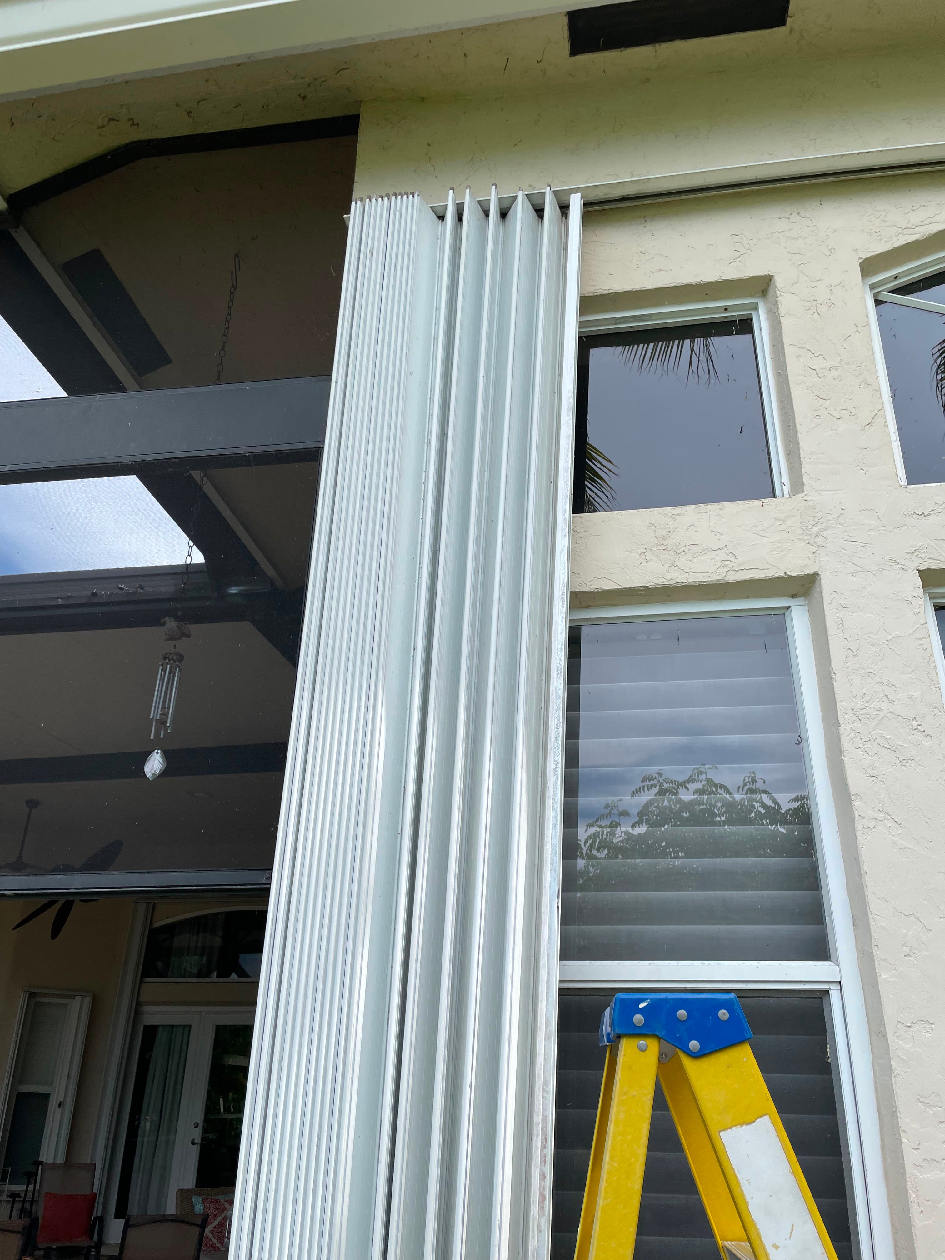 Accordion shutter stacked open against the jamb of a residential exterior wall beside windows, with a stepladder set up alongside the opening