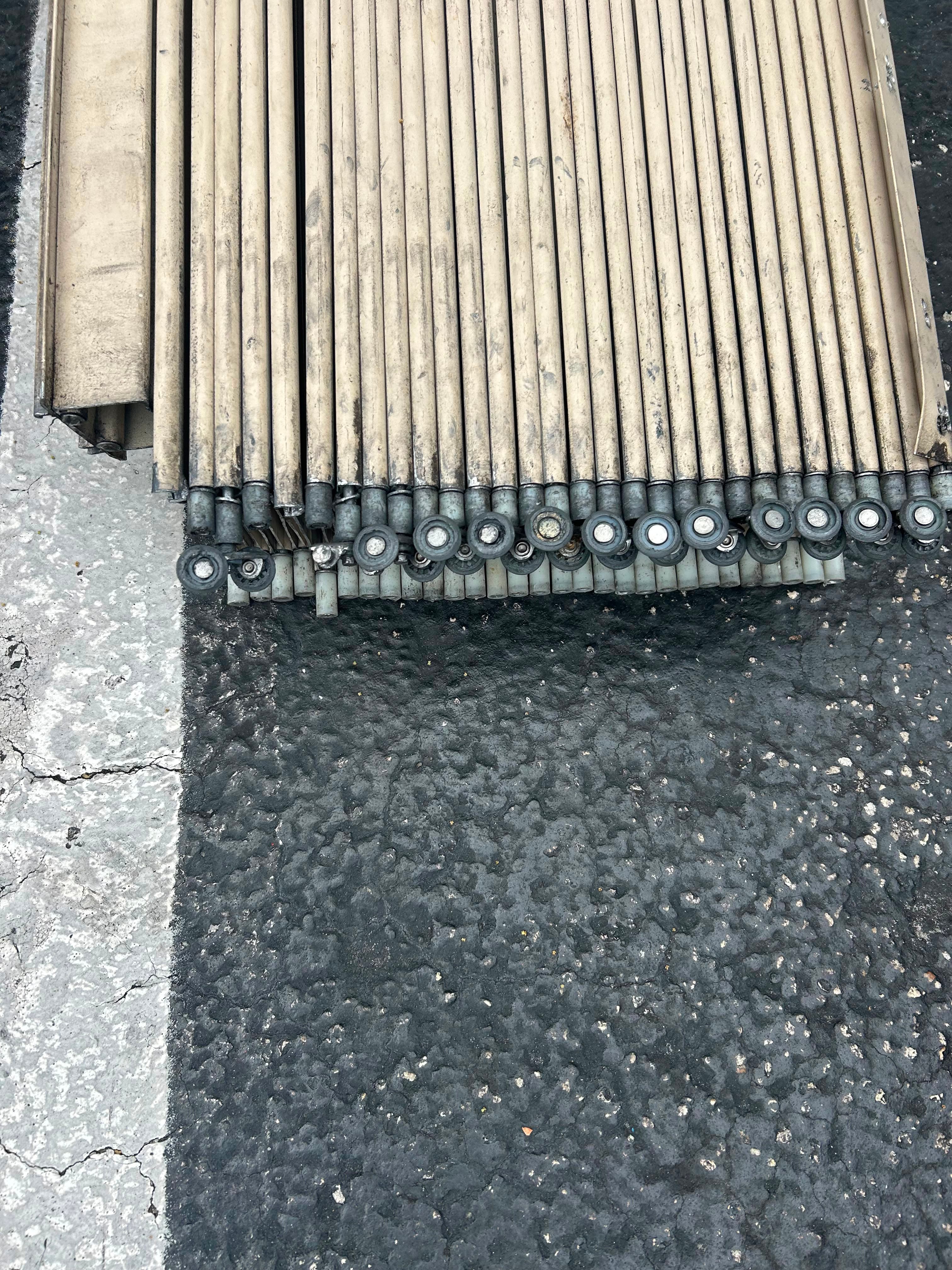 Close-up of the top edge of a removed accordion shutter curtain on asphalt, showing a row of weathered nylon wheel carriers and pivot pins