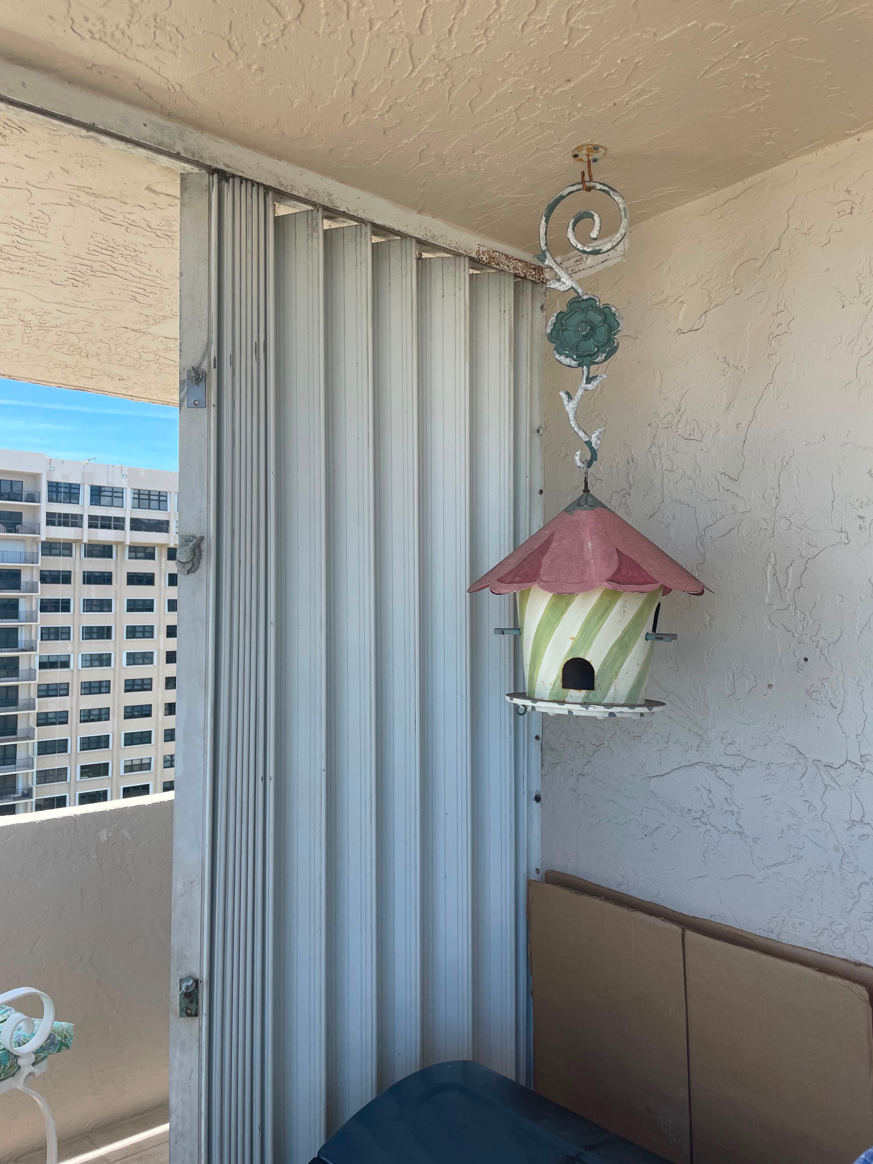 White accordion shutter partly drawn across a high-rise condominium balcony opening, with the upper track and mounting brackets visible above the folded blades