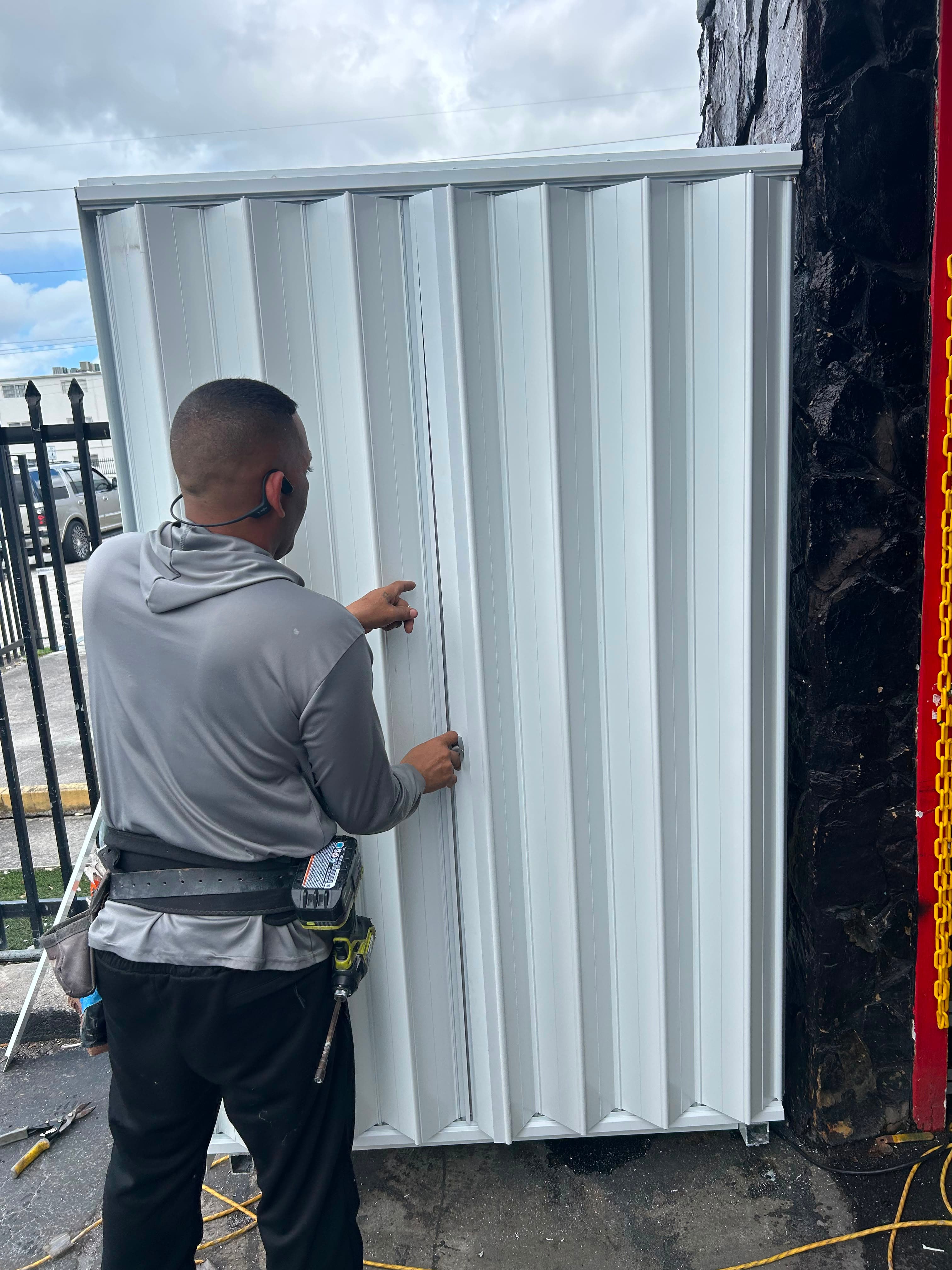 Technician with a tool belt and cordless drill working at the centre mating posts and lock of a closed white accordion shutter on a commercial storefront